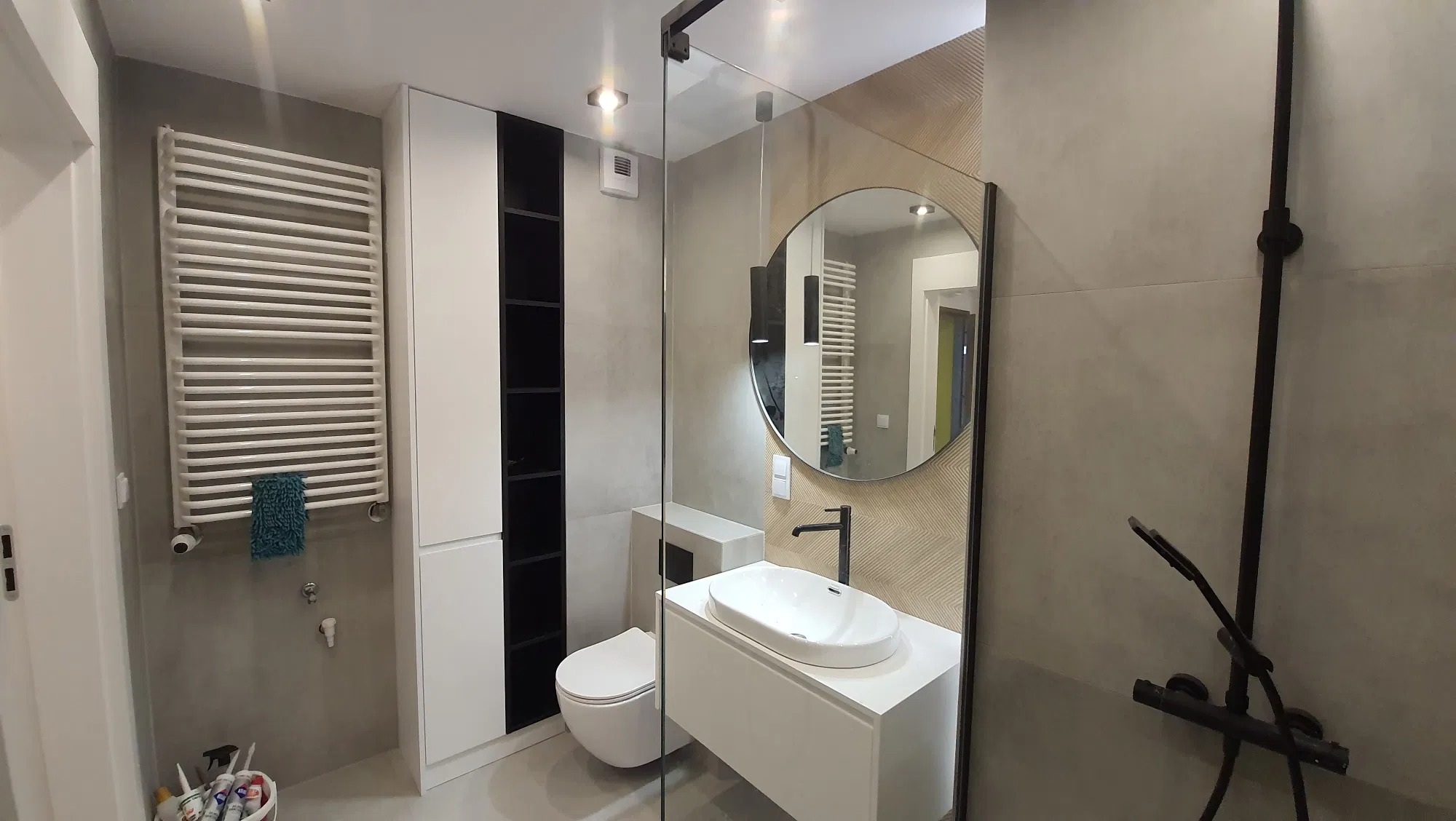 Nowoczesna łazienka z okrągłym lustrem, czarną armaturą i grzejnikiem drabinkowym, widoczna toaleta i kabina prysznicowa z czarnymi profilami.