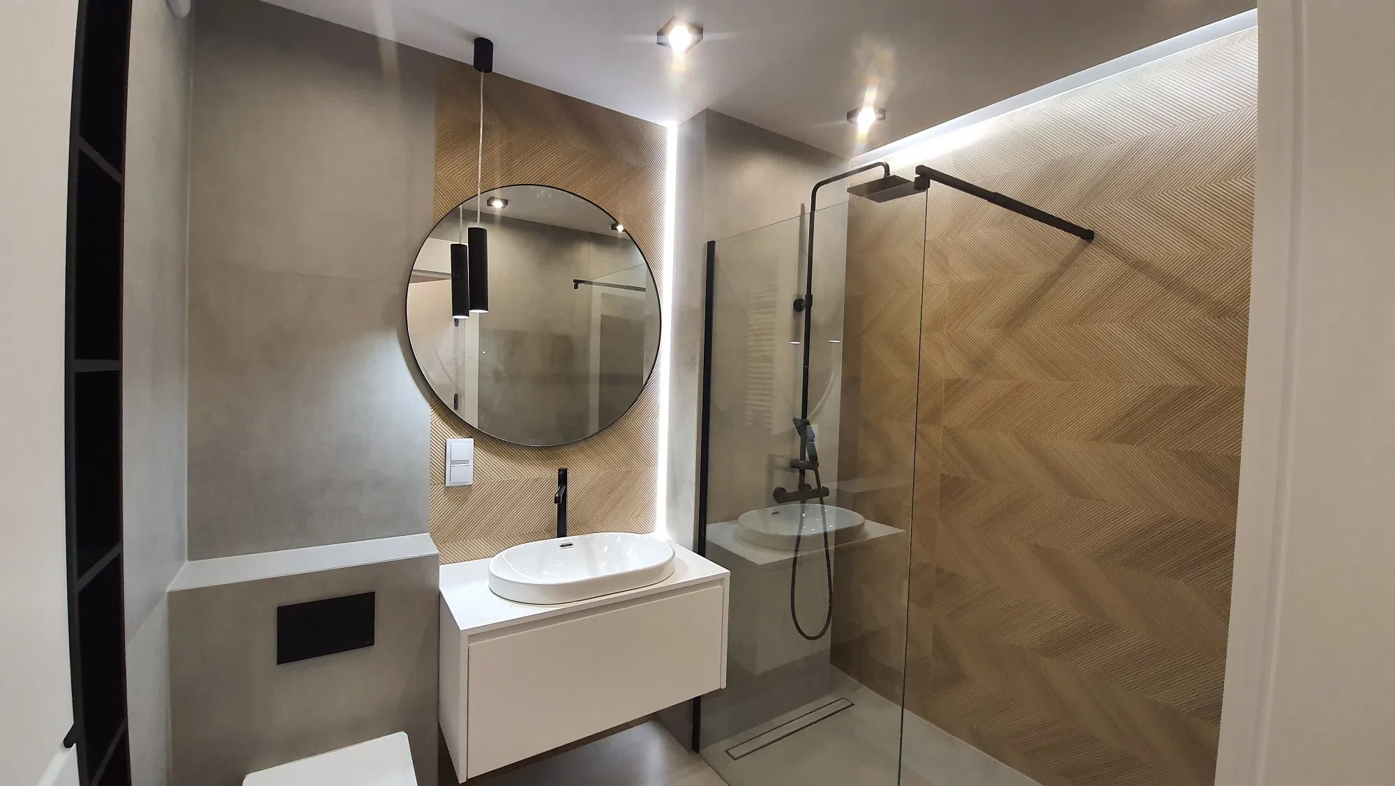 Nowoczesna łazienka z umywalką nablatową, okrągłym lustrem z podświetleniem LED, kabiną prysznicową typu walk-in i dekoracyjną ścianą z wzorem jodełki.