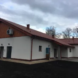 Budynek magazynowy z białymi ścianami, czerwoną dach&oacute;wką i drewnianą elewacją szczytową, widoczny klimatyzator na ścianie, otwarte drzwi wejściowe.