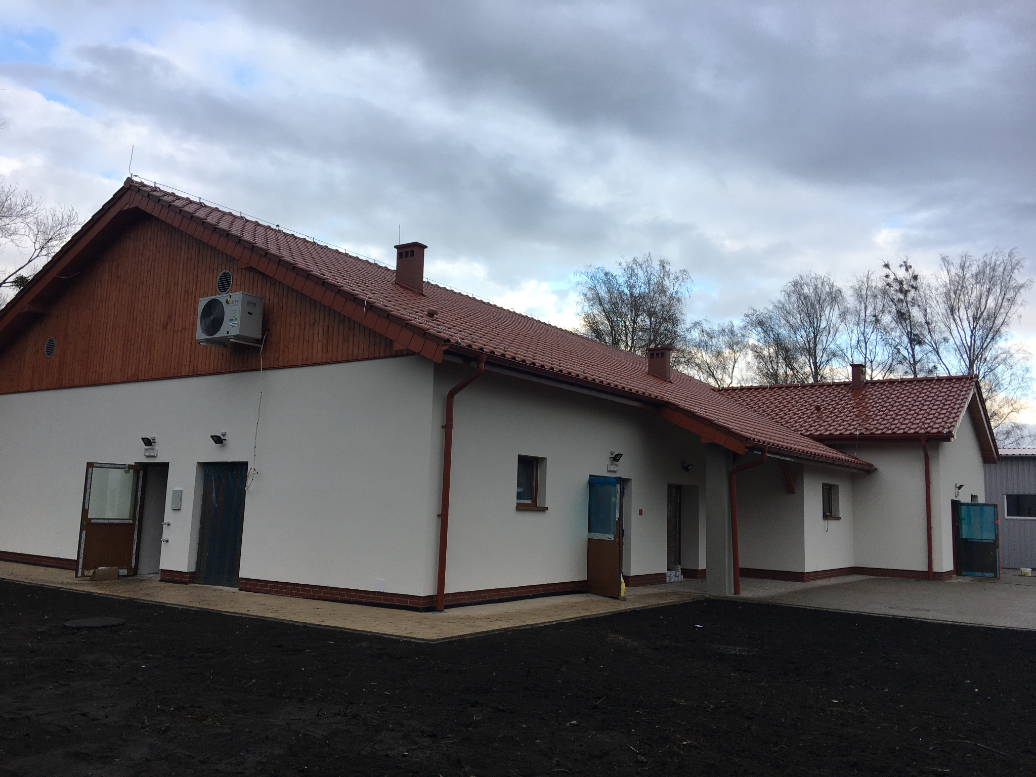 Budynek magazynowy z białymi ścianami, czerwoną dachówką i drewnianą elewacją szczytową, widoczny klimatyzator na ścianie, otwarte drzwi wejściowe.