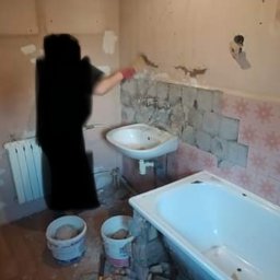 Tomasz Kubiak - Pracownik w czerwonej rękawicy nakłada zaprawę na ścianę w łazience z częściowo usuniętymi płytkami, widoczna wanna, umywalka, grzejnik i dwa wiadra z zaprawą oraz gruz na podłodze.