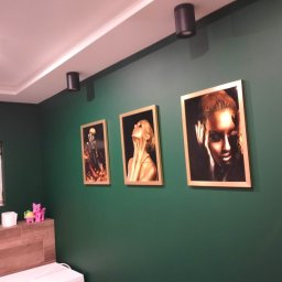 Tomasz Kubiak - Pomieszczenie z zielonymi ścianami, trzema obrazami w złotych ramach przedstawiającymi portrety kobiet, czarnymi lampami sufitowymi i fragmentem okna oraz półką z zabawkami.