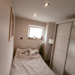 Tomasz Kubiak - Niewielka sypialnia w odcieniach beżu z łóżkiem przy ścianie, przesuwną szafą z lustrem i oknem dachowym z widokiem na niebo.