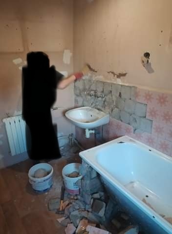 Pracownik w czerwonej rękawicy nakłada zaprawę na ścianę w łazience z częściowo usuniętymi płytkami, widoczna wanna, umywalka, grzejnik i dwa wiadra z zaprawą oraz gruz na podłodze.