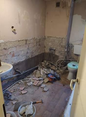 Łazienka w trakcie demontażu płytek i przygotowywania ścian do dalszych prac, widoczne odsłonięte fragmenty muru, gruz, rury, oraz częściowo zdemontowana toaleta z niebieską deską.