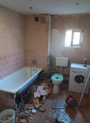 Łazienka w trakcie remontu: zdemontowane płytki, gruz, wanna, toaleta z zieloną deską, pralka i narzędzia na podłodze.