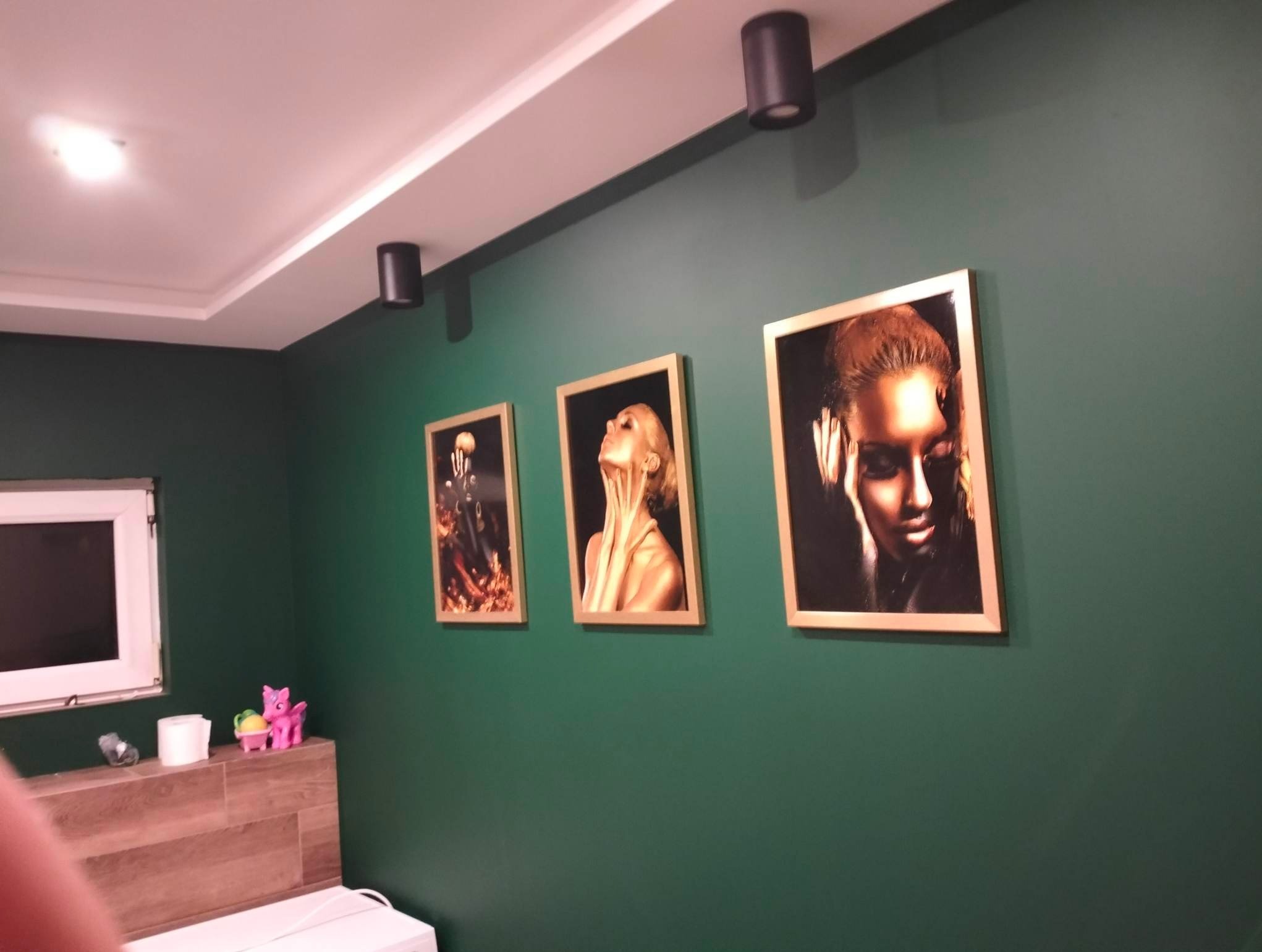 Pomieszczenie z zielonymi ścianami, trzema obrazami w złotych ramach przedstawiającymi portrety kobiet, czarnymi lampami sufitowymi i fragmentem okna oraz półką z zabawkami.