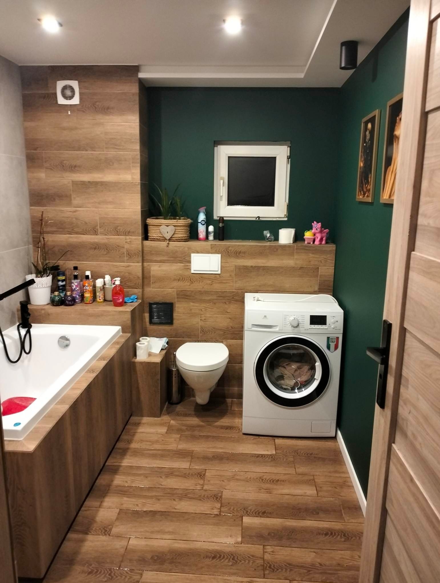 Ukończona łazienka z drewnopodobnymi płytkami na ścianach i podłodze, biała wanna obudowana drewnem, toaleta, pralka i ciemnozielona ściana z małym oknem.
