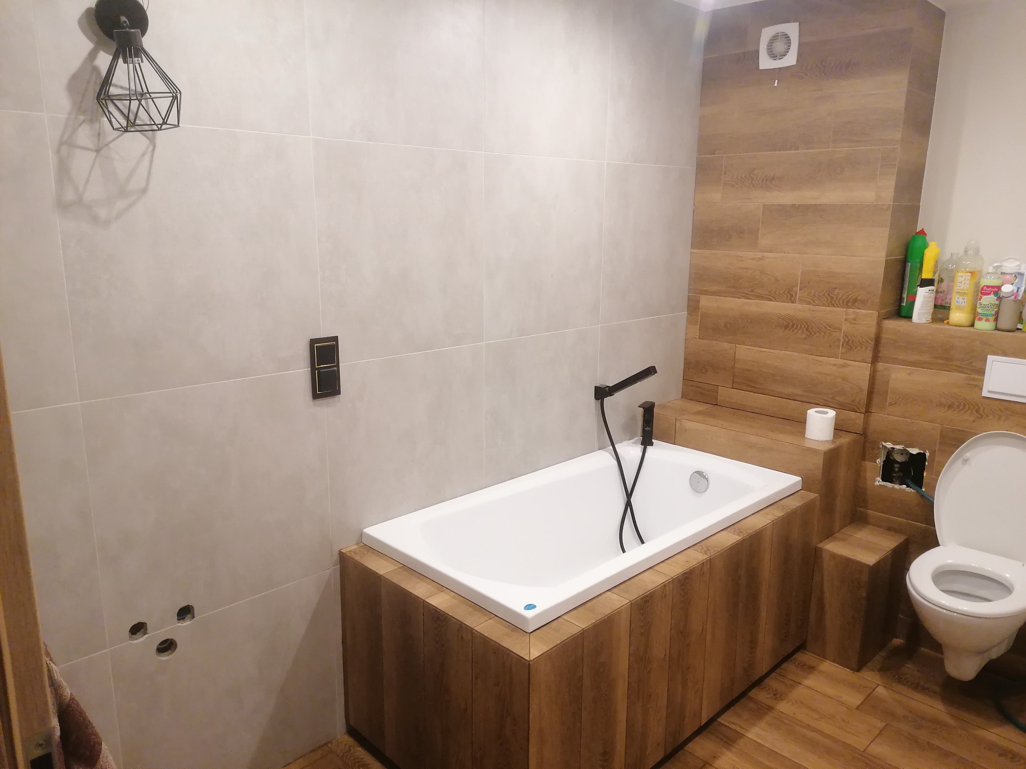 Nowoczesna łazienka z wanną obudowaną drewnopodobnymi panelami, czarną armaturą i toaletą, częściowo wykończona, widoczne otwory na kable i rury, oraz kosmetyki na półce.