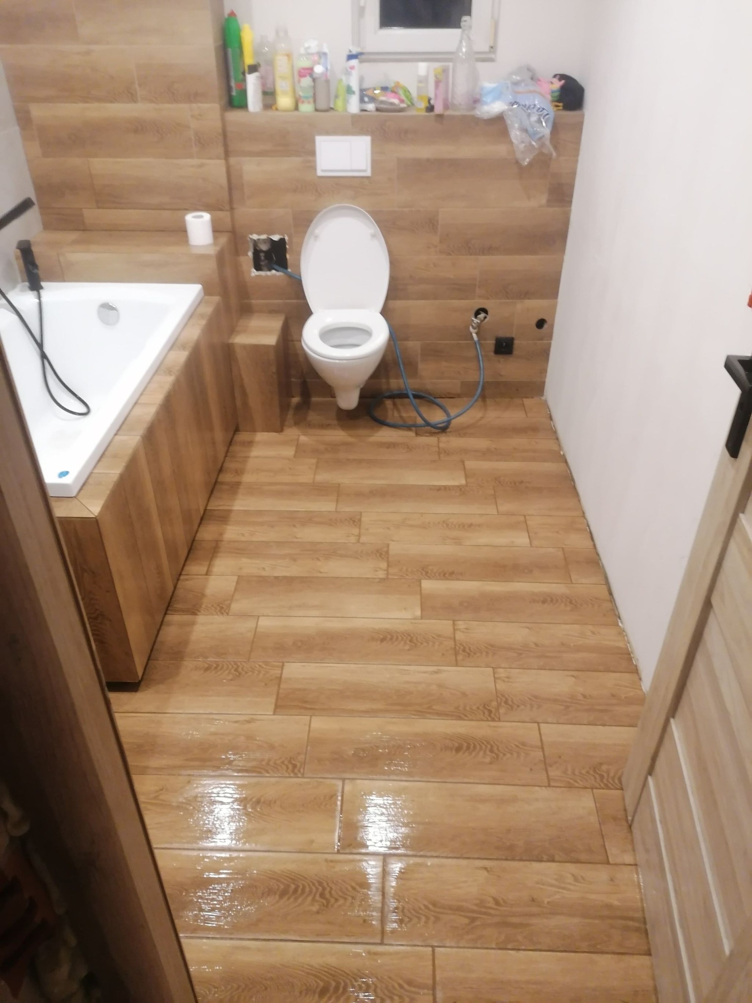 Ukończona łazienka z drewnopodobnymi płytkami na podłodze i ścianach, biała wanna obudowana panelami, wisząca toaleta i niezagruntowane miejsce po gniazdku.