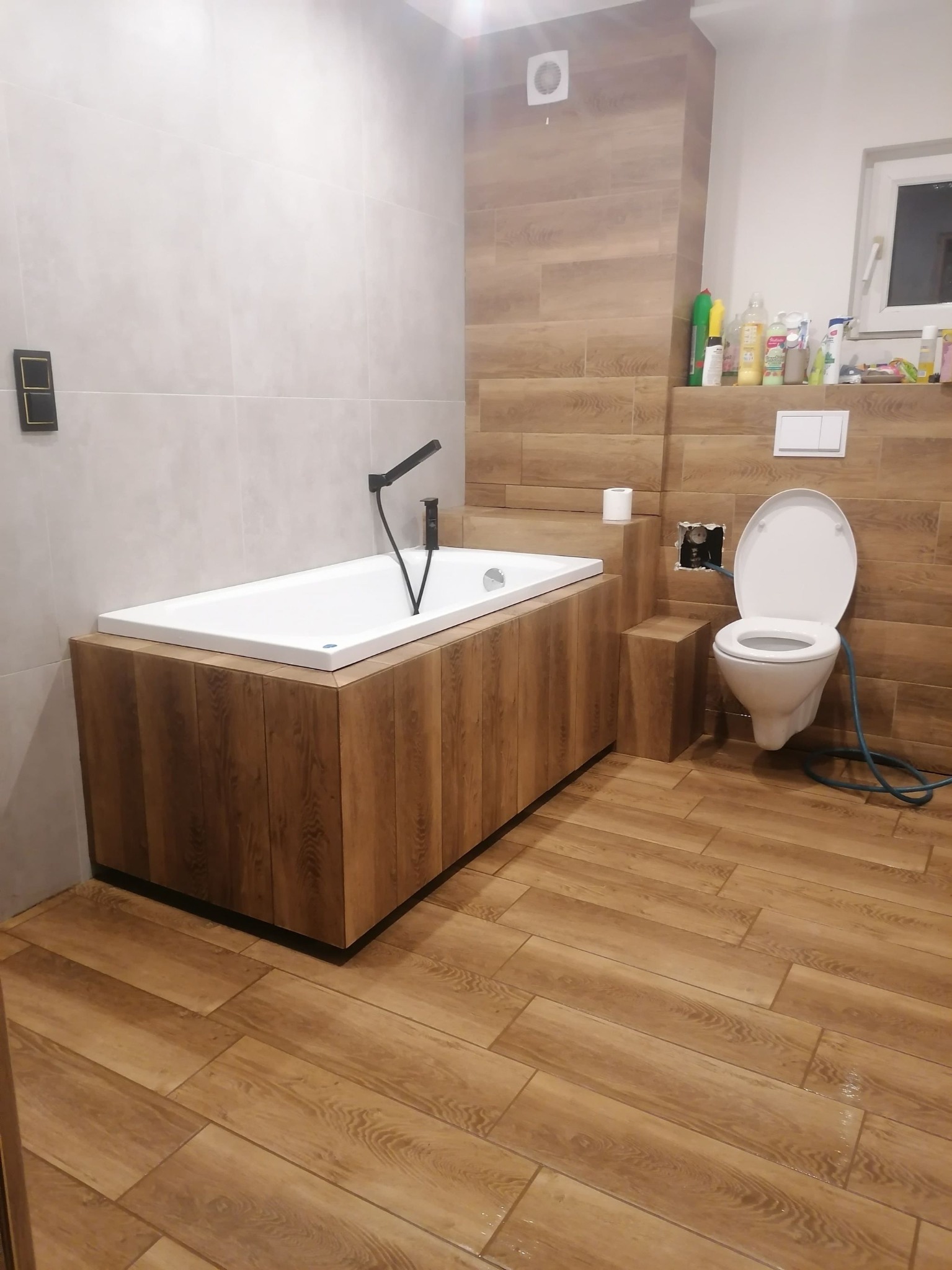 Wykończona łazienka z zabudowaną wanną z drewnopodobną obudową, podwieszaną toaletą z odsłoniętą instalacją, płytkami imitującymi drewno na podłodze i ścianie oraz białymi płytkami na ścianie za...