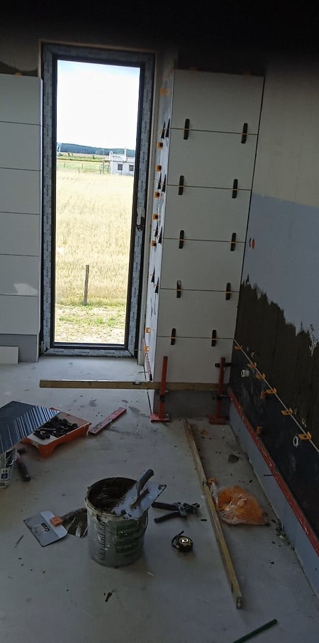 Wnętrze w trakcie kładzenia białych płytek ściennych z użyciem poziomicy laserowej i systemu poziomowania, widoczne narzędzia glazurnicze i wiadro z zaprawą, okno z widokiem na pole.