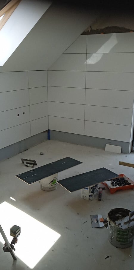 Łazienka na poddaszu w trakcie wykańczania, z ułożonymi białymi płytkami na ścianach, przygotowane ciemne płytki podłogowe leżą na wiadrach, widoczne narzędzia glazurnicze i poziomica laserowa.