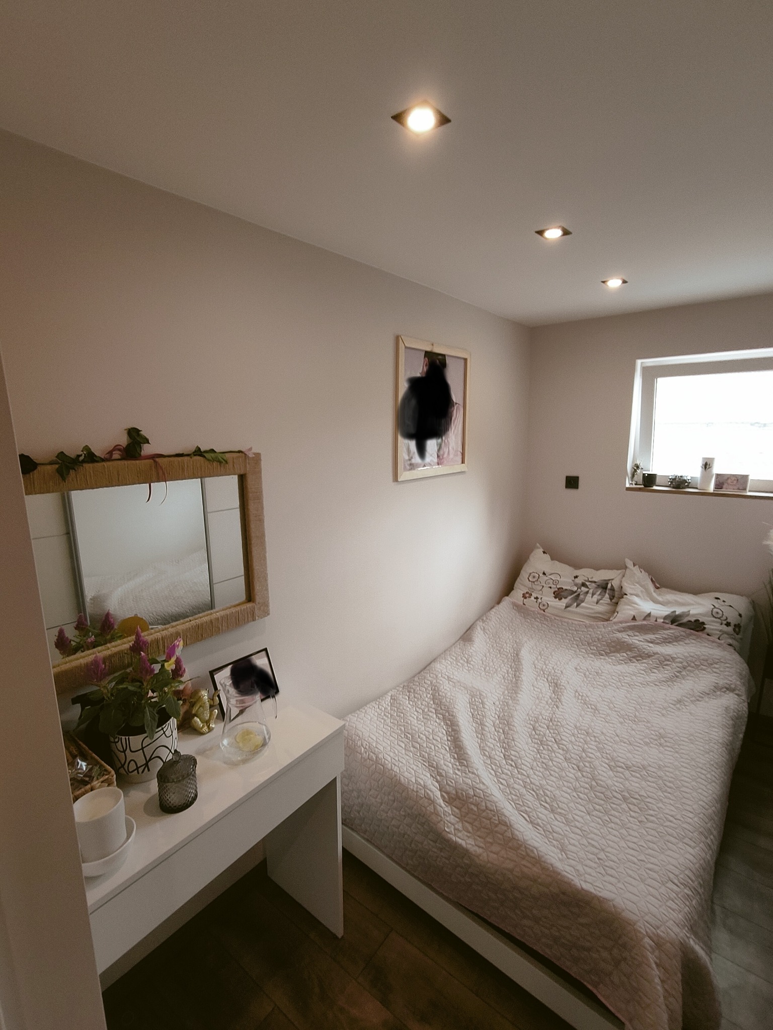 Jasna sypialnia z łóżkiem przy ścianie, toaletką z lustrem ozdobionym pnączami i oświetleniem punktowym w suficie.