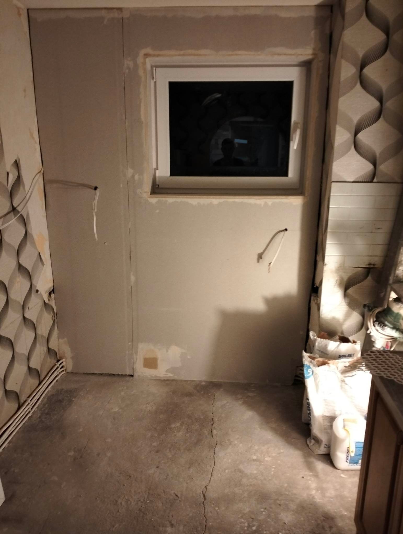 Łazienka w trakcie prac: ściany częściowo pokryte płytami gipsowo-kartonowymi, widoczne puszki elektryczne, okno z odbiciem osoby, worki z zaprawą murarską na podłodze.