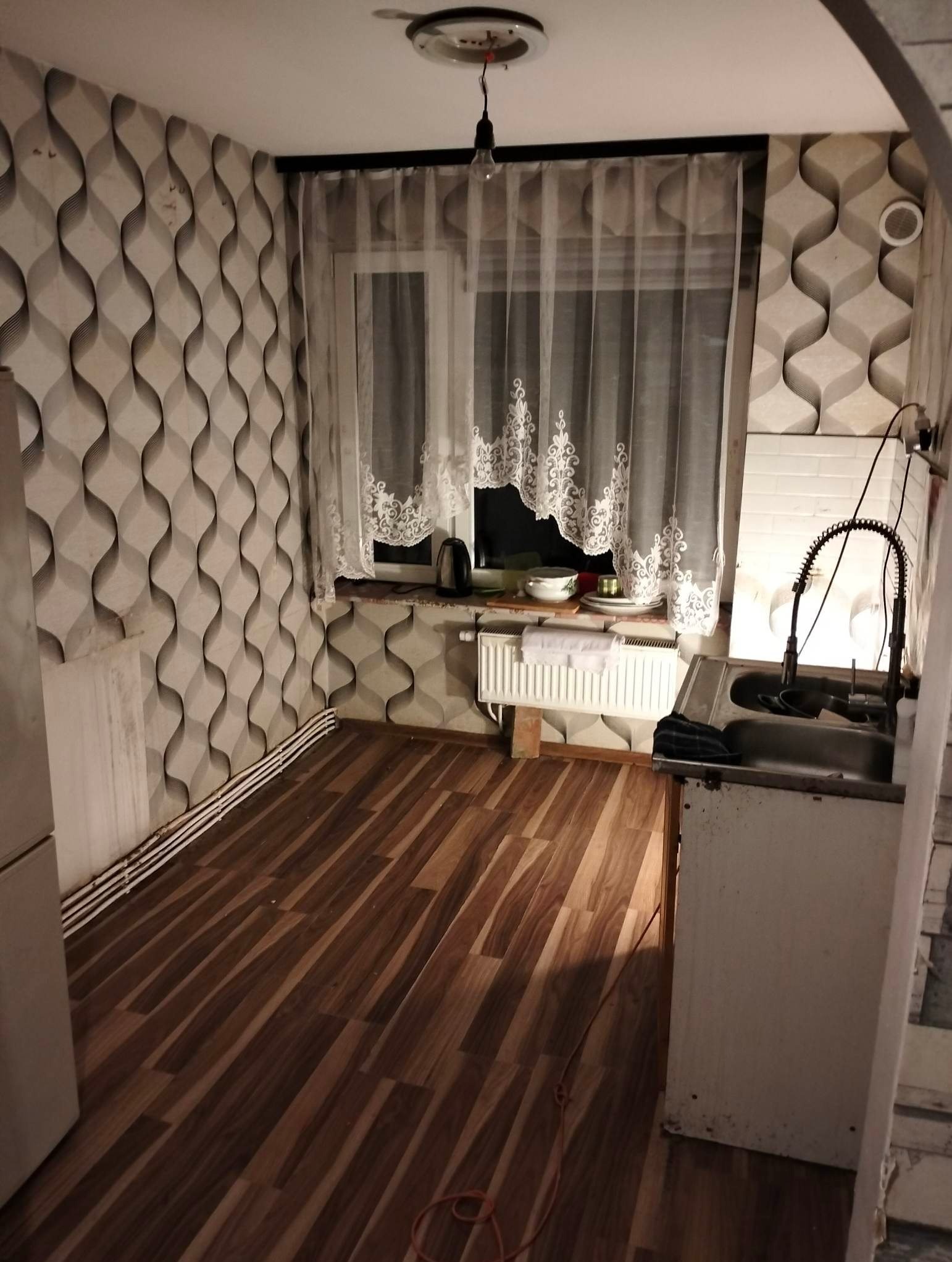 Wnętrze pokoju z drewnianą podłogą, wzorzystą tapetą, oknem z firanką i fragmentem blatu kuchennego z umywalką. Widoczny grzejnik pod oknem.