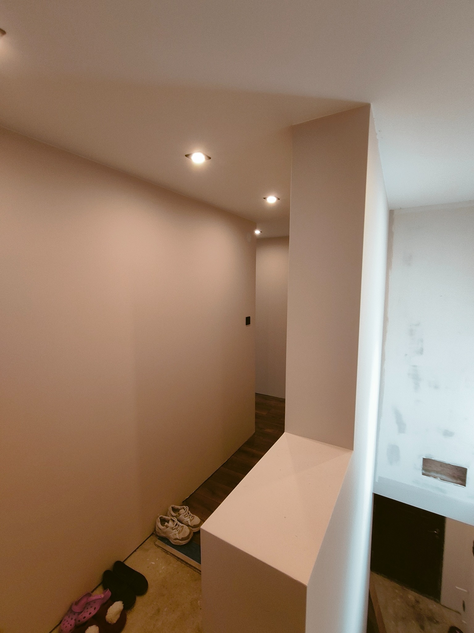 Korytarz w trakcie remontu z pomalowanymi ścianami na jasny kolor, widoczne buty i klapki na podłodze oraz wnęka z zabudową.