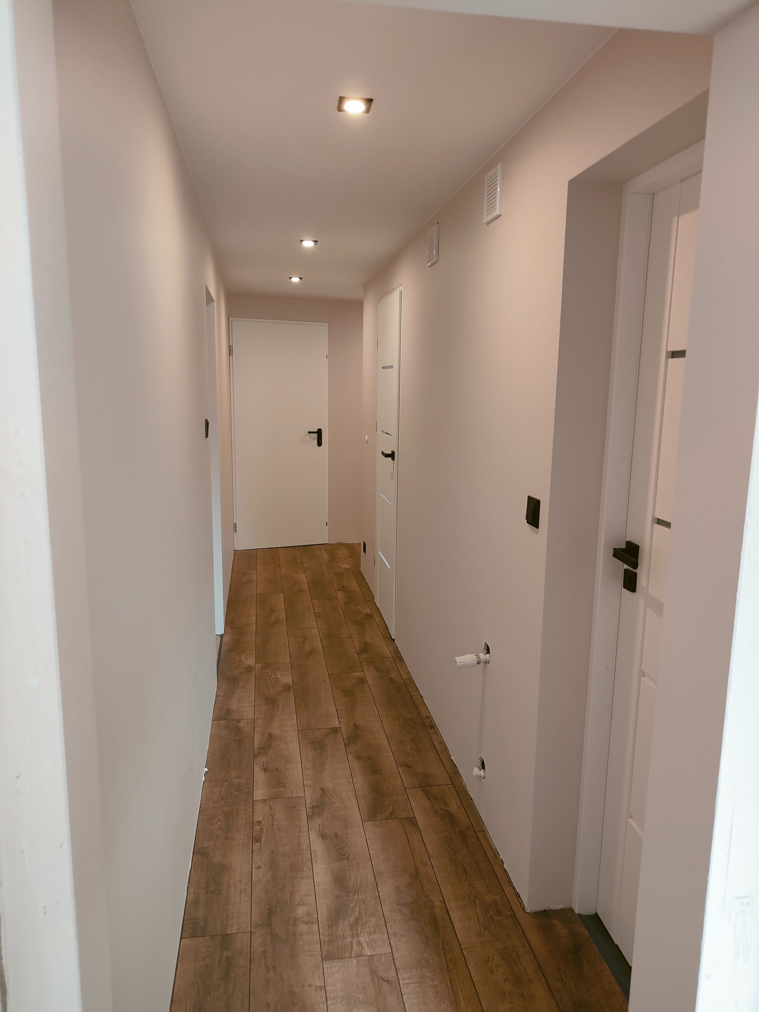 Długi korytarz po remoncie z panelami podłogowymi imitującymi drewno, białymi ścianami i białymi drzwiami z czarnymi klamkami. Widoczne punktowe oświetlenie sufitowe i wystające rury od grzejnika.