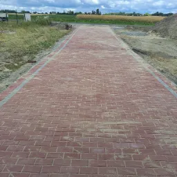 Nowa, czerwona kostka brukowa ułożona na długim podjeździe z widocznymi liniowymi odwodnieniami po bokach, otoczona trawnikiem i polem w tle pod pochmurnym niebem.