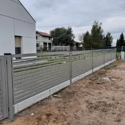 Nowoczesne, poziome ogrodzenie panelowe w kolorze szarym, osadzone na betonowej podmurówce, oddzielające posesję z nowoczesnym domem od drogi gruntowej.