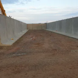 Wznoszenie ścian z prefabrykatów betonowych na placu budowy, widoczny dźwig i teren przygotowany pod wylewkę, stalowe pręty wystają z górnej krawędzi.