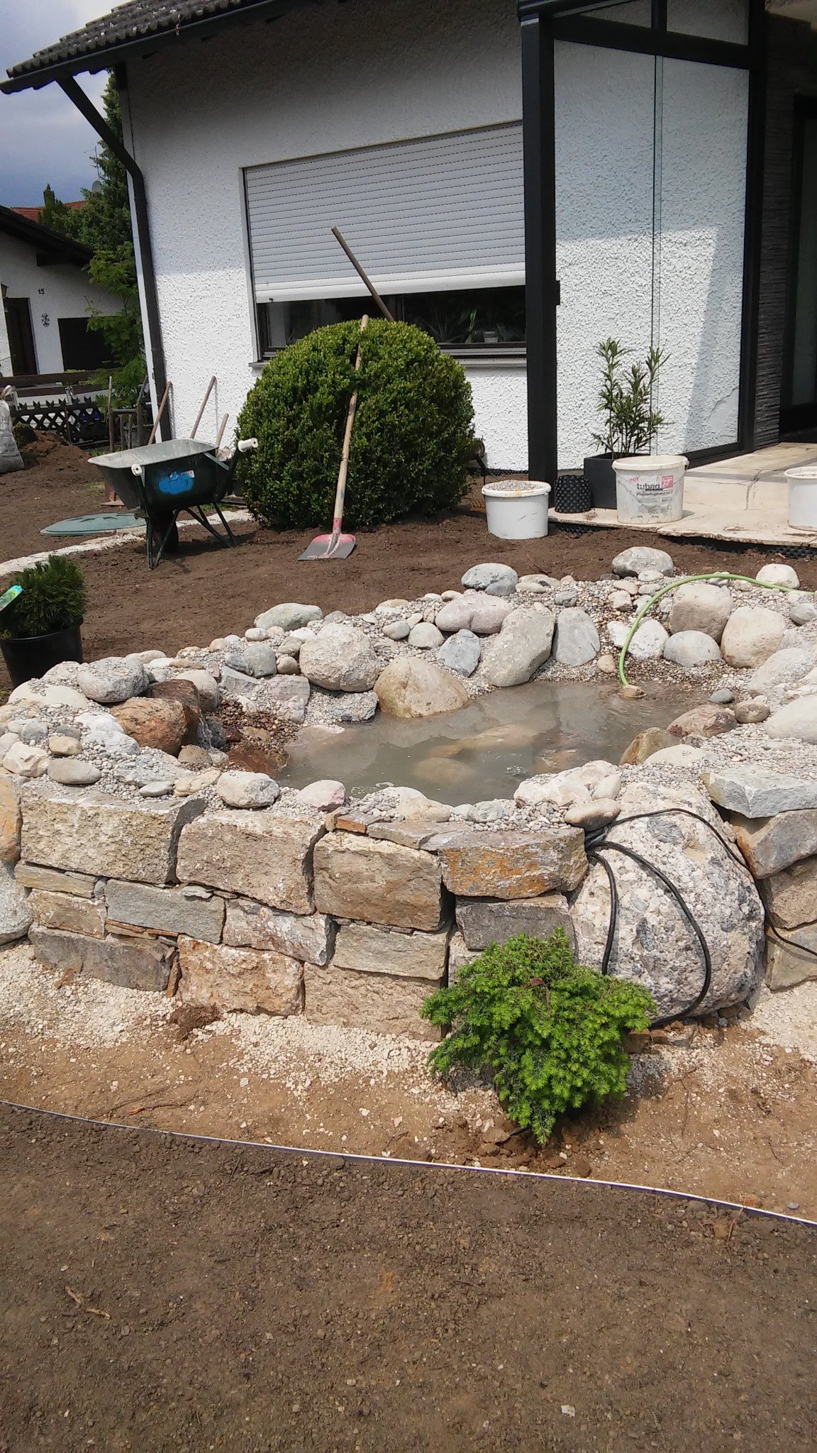 Budowa oczka wodnego z kamieni w ogrodzie, widoczna folia zabezpieczająca, taczka i narzędzia ogrodnicze w tle.