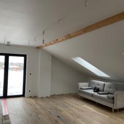 SK-SERVICES - Poddasze po remoncie z oknami dachowymi i drewnianą belką stropową, widoczna szara sofa, świeżo położona podłoga i przygotowane punkty oświetleniowe.