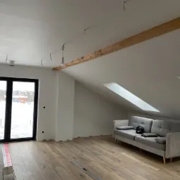 Poddasze po remoncie z oknami dachowymi i drewnianą belką stropową, widoczna szara sofa, świeżo położona podłoga i przygotowane punkty oświetleniowe.
