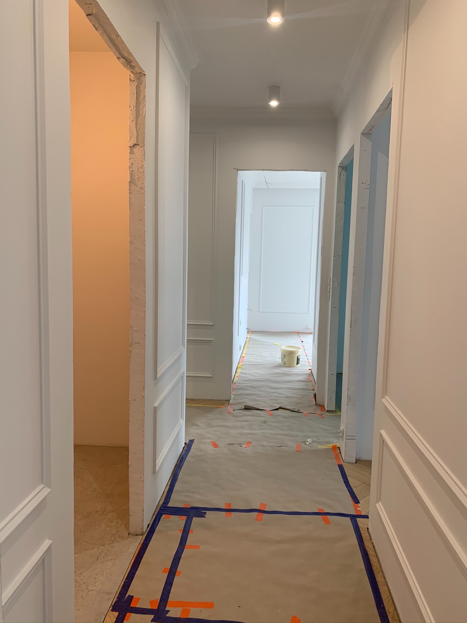 Korytarz w trakcie prac wykończeniowych, zabezpieczona podłoga brązowym papierem i niebieską taśmą, widoczne ściany z białymi panelami i otwarte przejścia do innych pomieszczeń, surowy otwór...