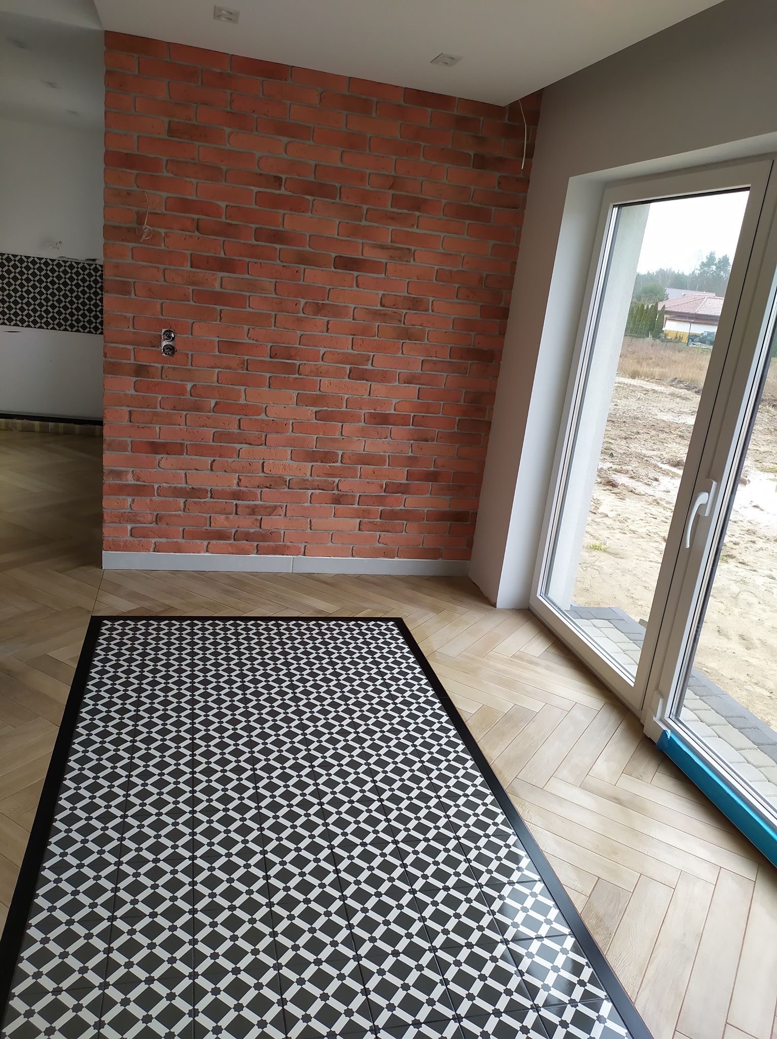 Wykończone wnętrze z ceglaną ścianą, podłogą w jodełkę i czarno-białą mozaiką na posadzce, widok na zewnątrz przez duże okno.