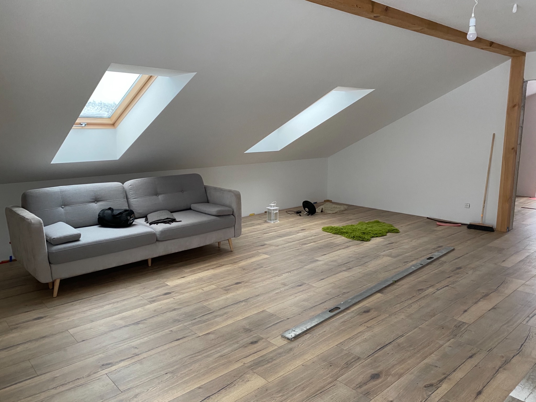 Poddasze z drewnopodobną podłogą, szarym narożnikiem i dwoma oknami dachowymi, poziomica leżąca na podłodze, widoczny fragment drewnianej belki stropowej.