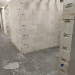 Wnętrze w trakcie budowy, widać wznoszone ściany z białych bloczków betonowych, z drewnianymi belkami stropowymi mocowanymi metalowymi łącznikami, na podłodze widoczne ślady zaprawy.