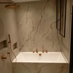 Nowoczesna łazienka z białą wanną, marmurowymi płytkami, złotą armaturą i czarnym grzejnikiem drabinkowym.