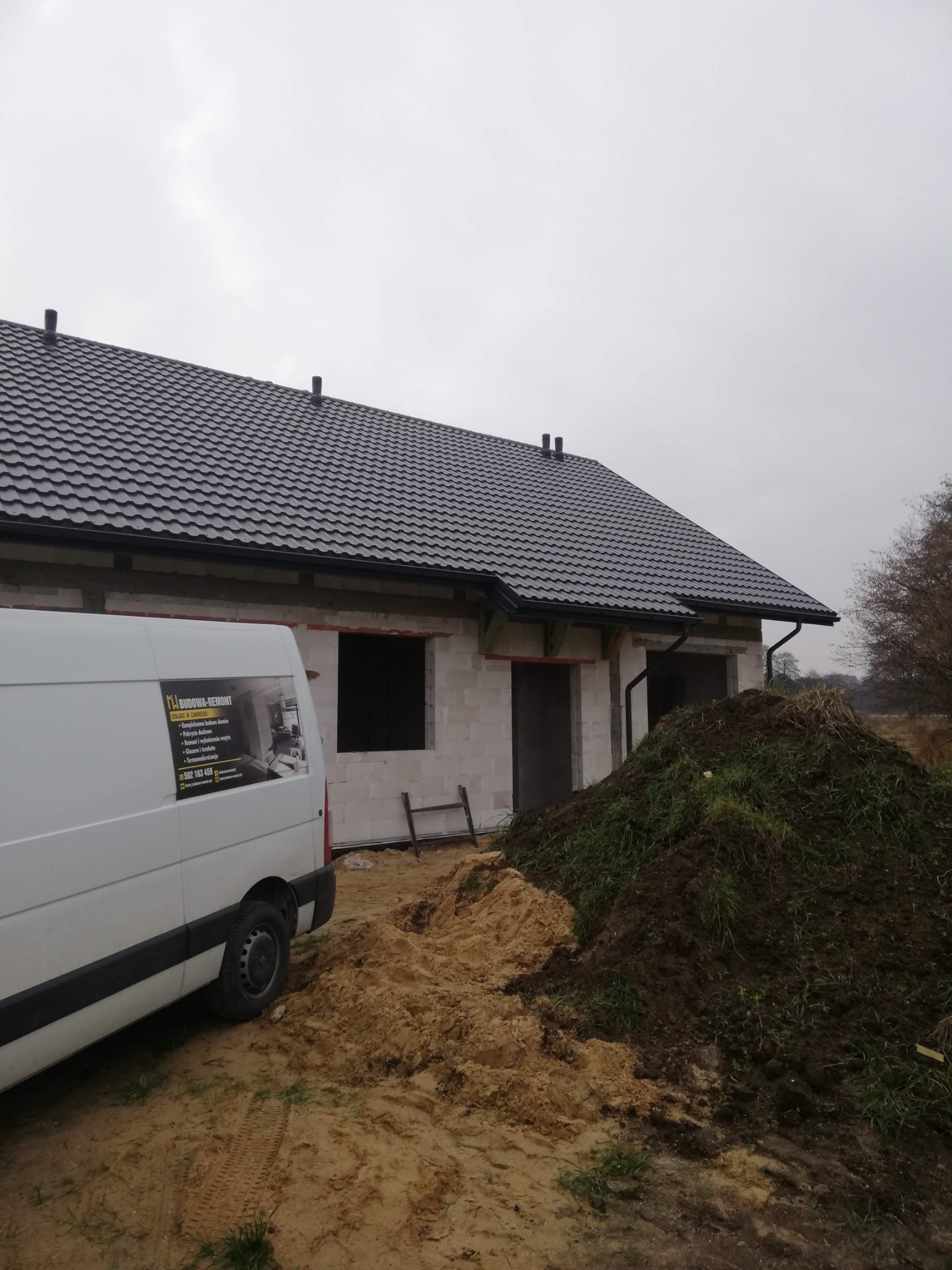 Niedokończony budynek jednorodzinny z ciemną dachówką, widoczny z boku, z zaparkowanym białym busem firmy budowlanej i stertą ziemi na pierwszym planie.