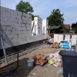 Budowa domu z bloczków betonu komórkowego: murarz pracuje przy wznoszeniu ściany, widoczne bloczki Silka na paletach, izolacja pozioma i stos cegieł klinkierowych. Śmieci w czarnym koszu...