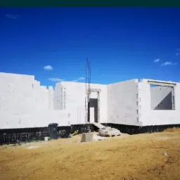 Wznoszenie ścian parterowego domu z betonu komórkowego, widoczne zbrojenie pionowe i hydroizolacja fundamentów, w tle błękitne niebo.