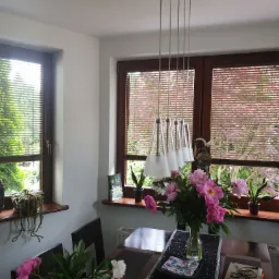 Jadalnia z dwoma oknami, na których zamontowano poziome żaluzje w kolorze drewna. Na parapetach dekoracje i kwiaty. Na stole wazon z piwoniami.