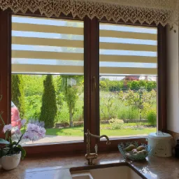 Widok z okna kuchennego na ogród przez poziome pasy beżowo-białej osłony okiennej, firanka z koronki u góry, kran w stylu retro, kwiat storczyka w doniczce na blacie.