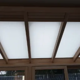 Białe, plisowane osłony przeciwsłoneczne zamontowane pod drewnianą konstrukcją dachu werandy, widok z dołu.