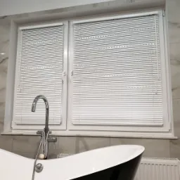 Białe żaluzje poziome zamontowane w oknie nad czarno-białą wanną w łazience z marmurowymi płytkami.