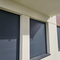 FHU Darpol Dariusz Rypuła - Elewacja budynku z trzema oknami zabezpieczonymi szarymi roletami zewnętrznymi, widoczne fragmenty ceglanej dekoracji i ogrodzenia.