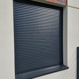 FHU Darpol Dariusz Rypuła - Zamknięta, grafitowa roleta zewnętrzna na oknie budynku z elewacją w jasnym odcieniu i fragmentem muru z cegły. Widoczne poziome lamele rolety i parapet.
