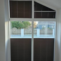 FHU Darpol Dariusz Rypuła - Ciemnobrązowe plisy okienne zamontowane w oknie o nieregularnym kształcie. Widok na zewnątrz, gdzie widać ogrodzenie i dom. Plisy dopasowane do nietypowego kształtu okna.