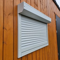 FHU Darpol Dariusz Rypuła - Biała roleta zewnętrzna częściowo opuszczona na oknie drewnianego budynku. Widoczne poziome lamele i kaseta rolety. Ujęcie z boku, podkreślające detale montażu.