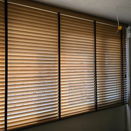 FHU Darpol Dariusz Rypuła - Zamknięte żaluzje drewniane w ciepłym odcieniu brązu, z czarnymi sznurkami, zamontowane w oknie. Widoczne elementy sufitu i ściany w neutralnych kolorach.