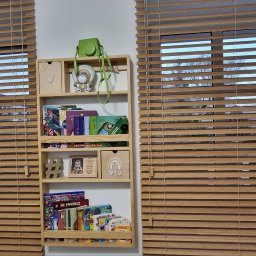 FHU Darpol Dariusz Rypuła - Drewniane żaluzje poziome w ciepłym odcieniu drewna, zamontowane w oknach pokoju dziecięcego, z półką na książki i zabawki pośrodku. Naturalne światło dzienne.