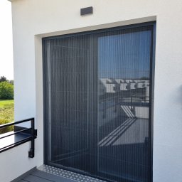 FHU Darpol Dariusz Rypuła - Zamknięta, plisowana moskitiera w grafitowej ramie, zamontowana na balkonie z szarą podłogą. Widok z zewnątrz, w tle zabudowa i zieleń.