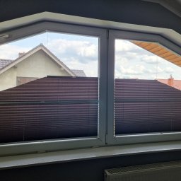 FHU Darpol Dariusz Rypuła - Brązowe plisy w oknie o nietypowym kształcie, widok na dom i niebo. Plisy częściowo zasłonięte, z widoczną strukturą materiału.