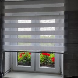 FHU Darpol Dariusz Rypuła - Okno z białą ramą i zamontowaną roletą dzień noc, przez którą widać czerwone kwiaty na zewnątrz. Fragment ściany z szaro-białymi płytkami w tle.