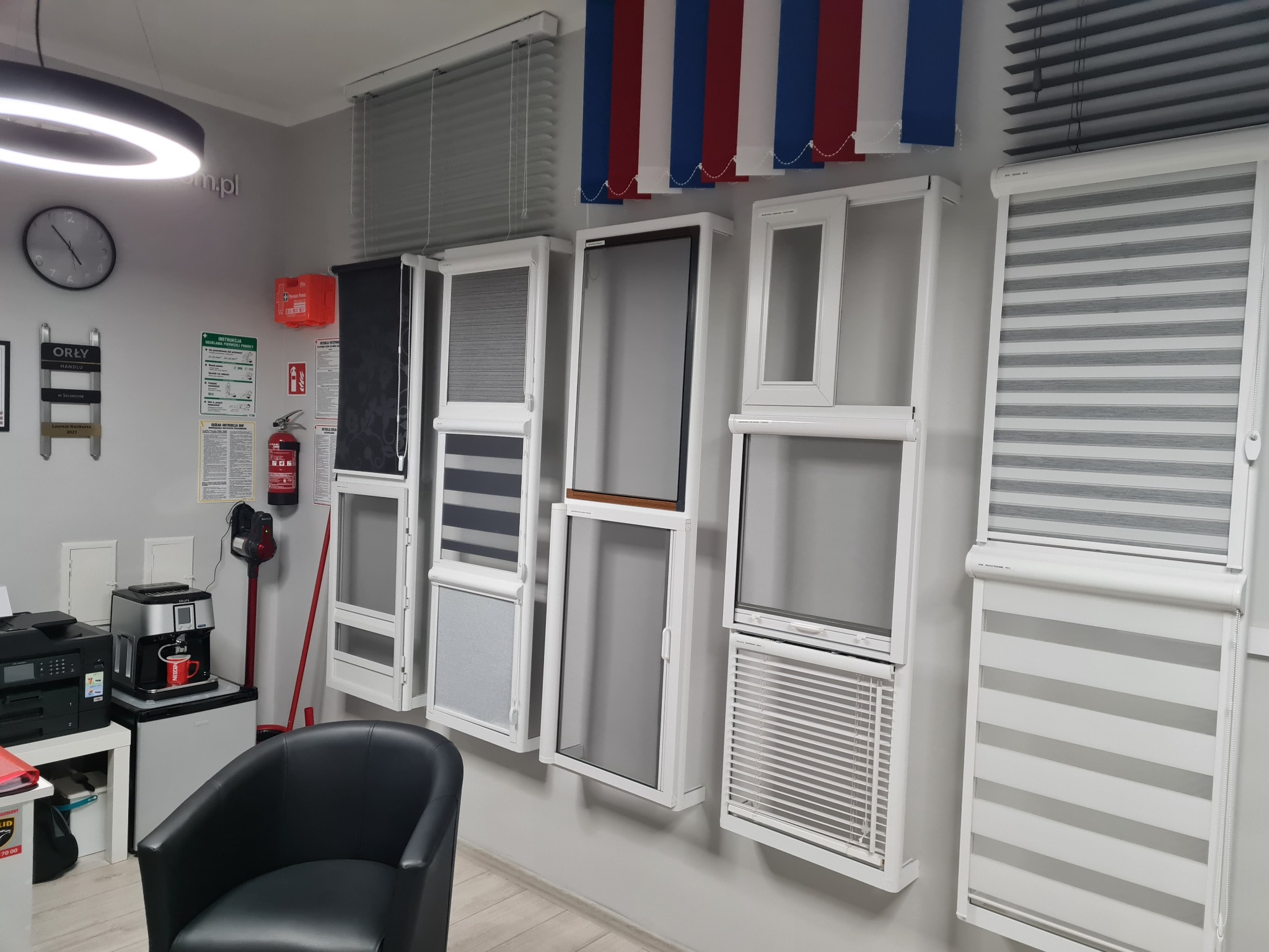 Ekspozycja różnych typów rolet i moskitier w salonie: rolety dzień-noc, plisy, żaluzje aluminiowe i drewniane, moskitiery ramkowe w białych ramach. Prezentacja asortymentu.